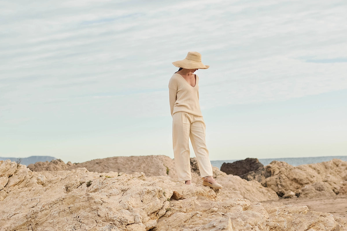 A person wearing a beige knit long-sleeve top and matching wide-leg pants stands on rocky terrain outdoors under a pale sky, with a wide-brim straw hat in hand.