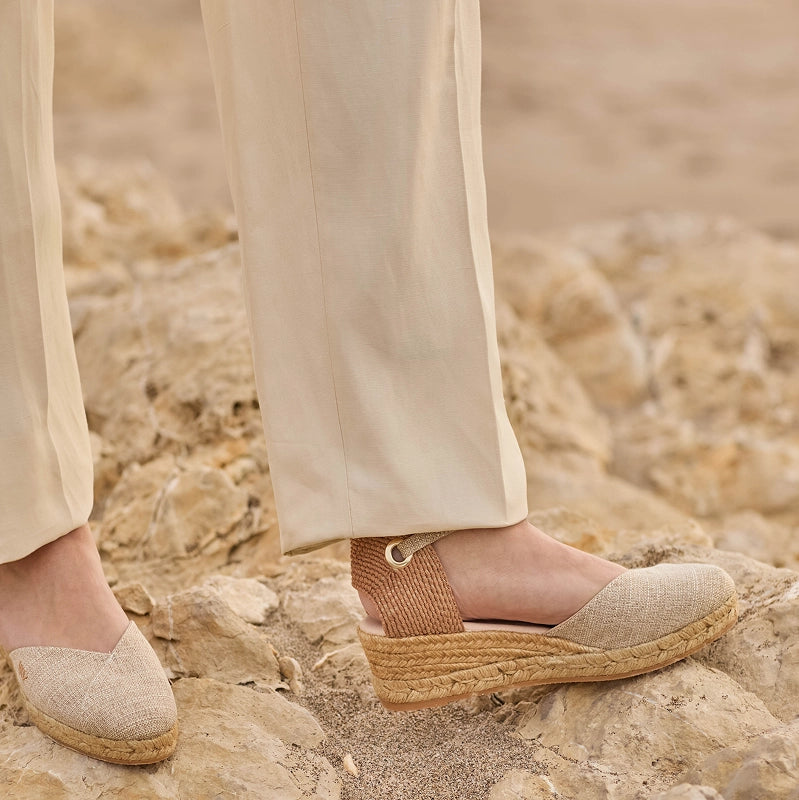 A close-up of a person wearing beige trousers and a pair of espadrille wedge sandals with woven jute soles and ankle straps, standing on a rocky, sandy surface.