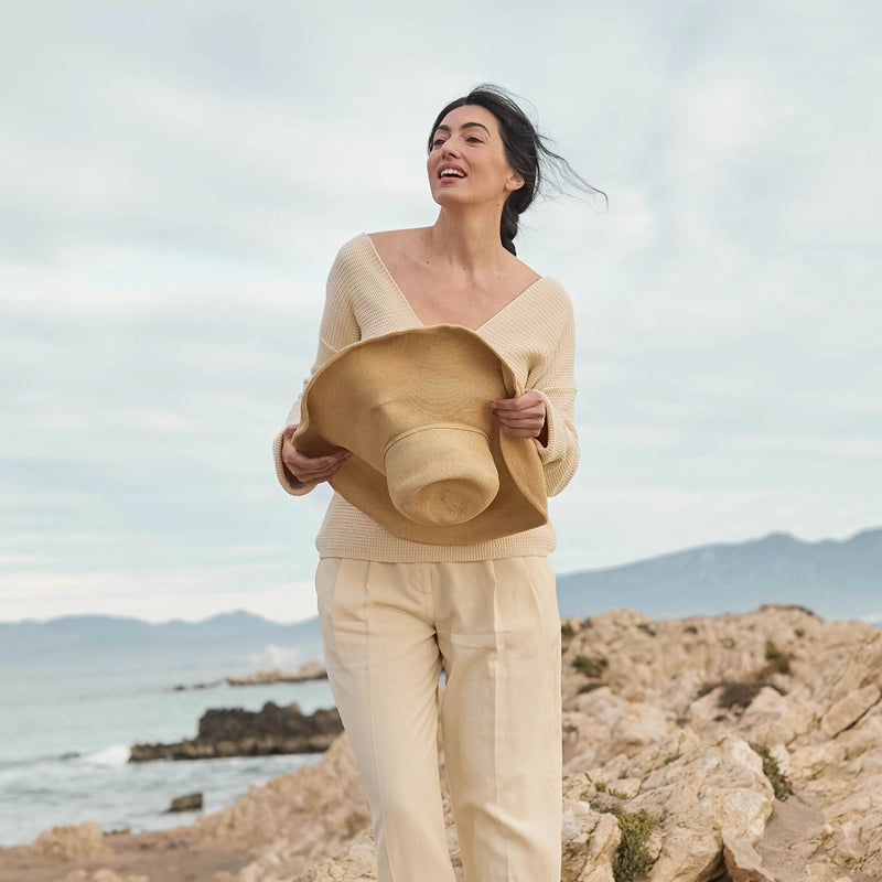 A woman stands on a rocky coastal path wearing a cream ribbed knit V-neck sweater and matching cream pants, holding a wide-brimmed straw hat. The background shows the ocean, distant mountains, and a cloudy sky.