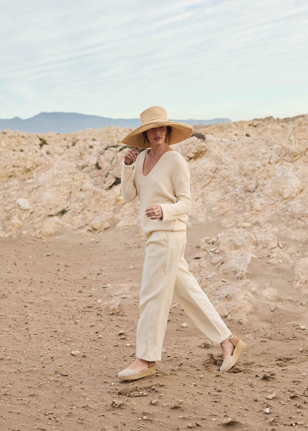 Woman walking along a sandy coastal path under soft morning light, wearing Pubol gold canvas ankle-strap wedge espadrilles with light cream trousers, a textured sweater, and a wide straw hat — a look of natural elegance and quiet confidence.