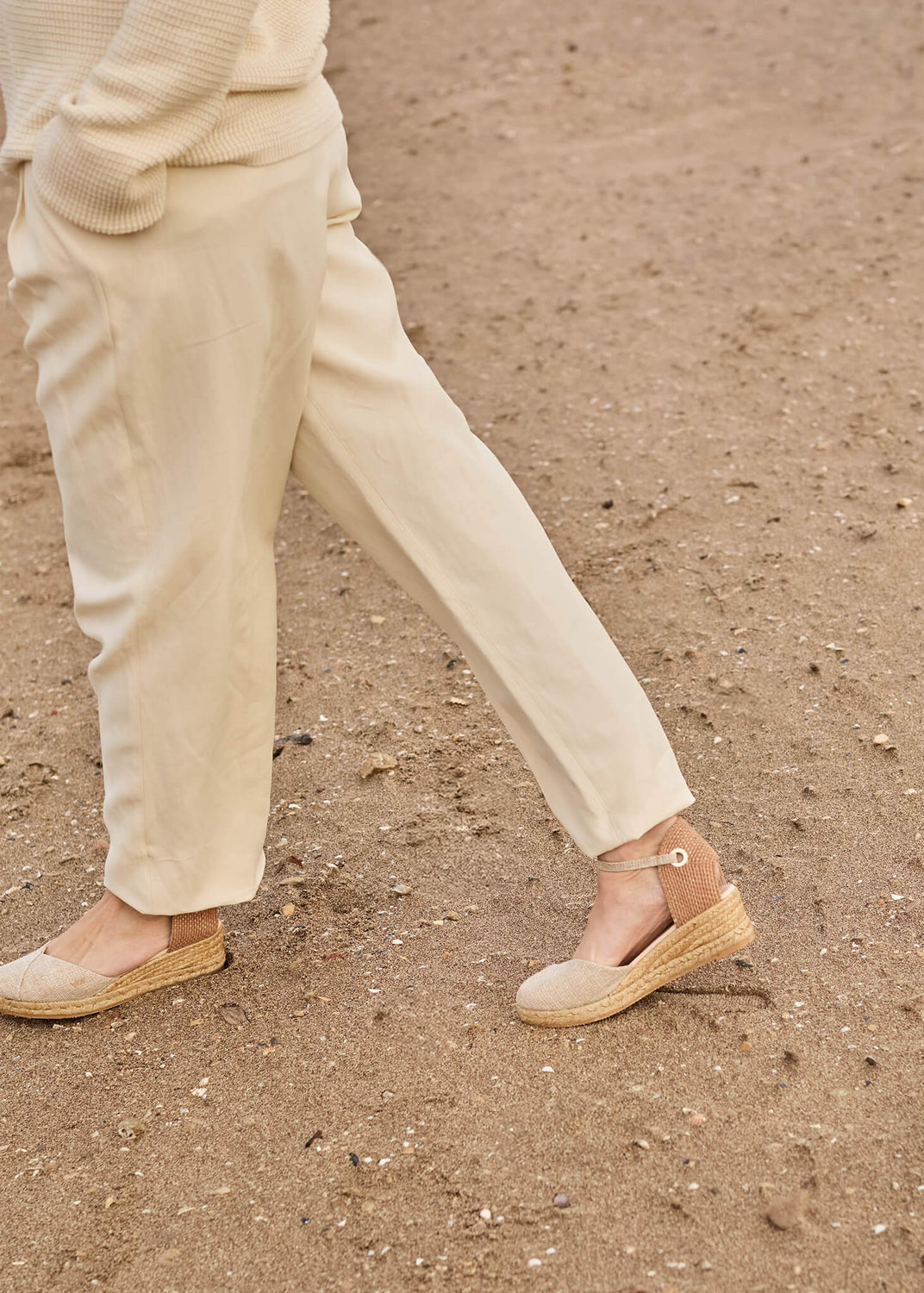 Close-up of a woman walking on soft sand, dressed in light beige trousers and the Pubol Gold wedge espadrilles. The look captures effortless movement and the soft glow of Mediterranean simplicity.