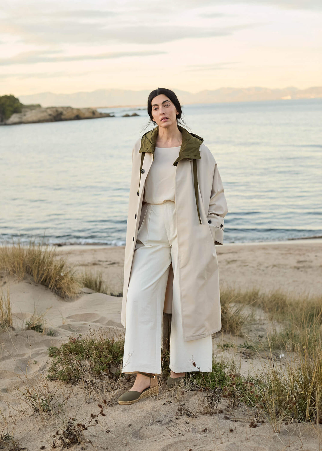 Woman standing on a sandy beach at sunset wearing Pubol olive canvas ankle-strap wedge espadrilles, light cream trousers, and a beige trench coat, evoking effortless coastal elegance.