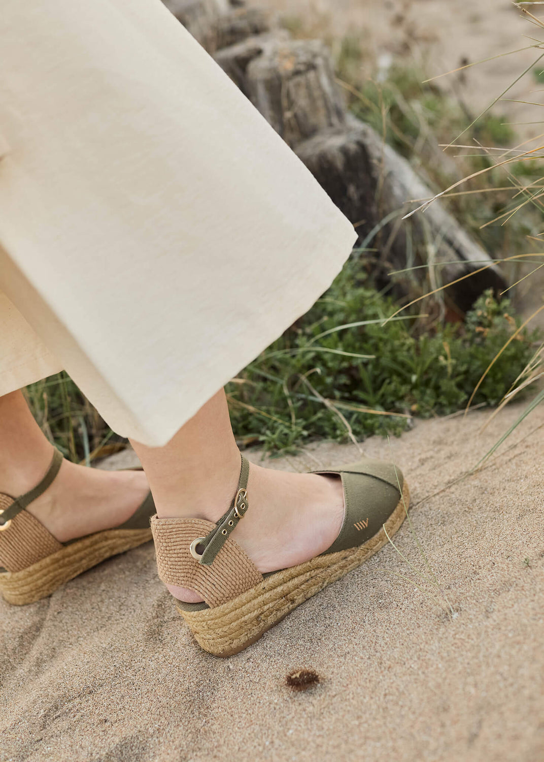 Close-up of a woman wearing Pubol olive canvas ankle-strap wedge espadrilles with a light cream skirt, walking barefoot on a sunlit sandy path surrounded by coastal greenery.