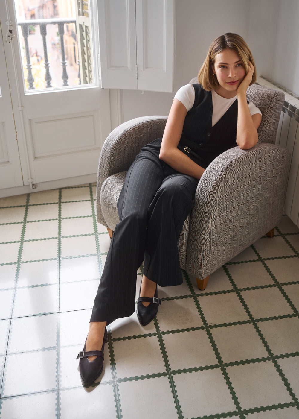 Woman sitting on a gray armchair wearing a white top, black vest, and pinstripe trousers with black leather flats featuring buckle detail — Viscata Remini shoes lifestyle image.
