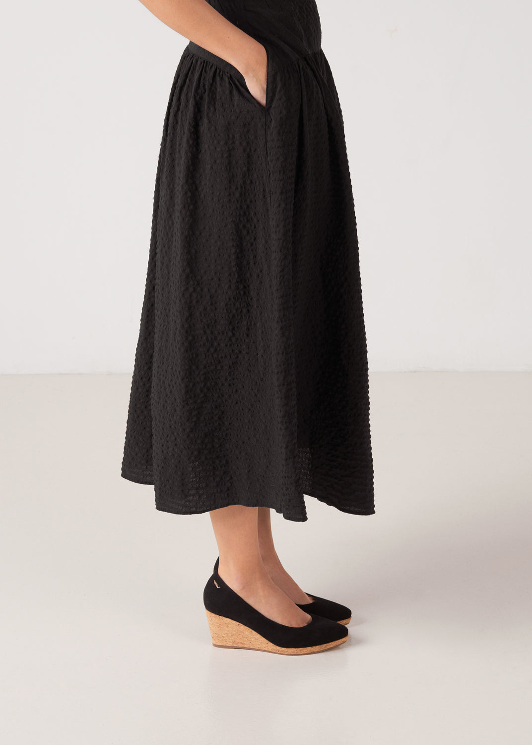 Side view of black suede slip-on wedge shoes with a cork sole, styled with a black midi dress