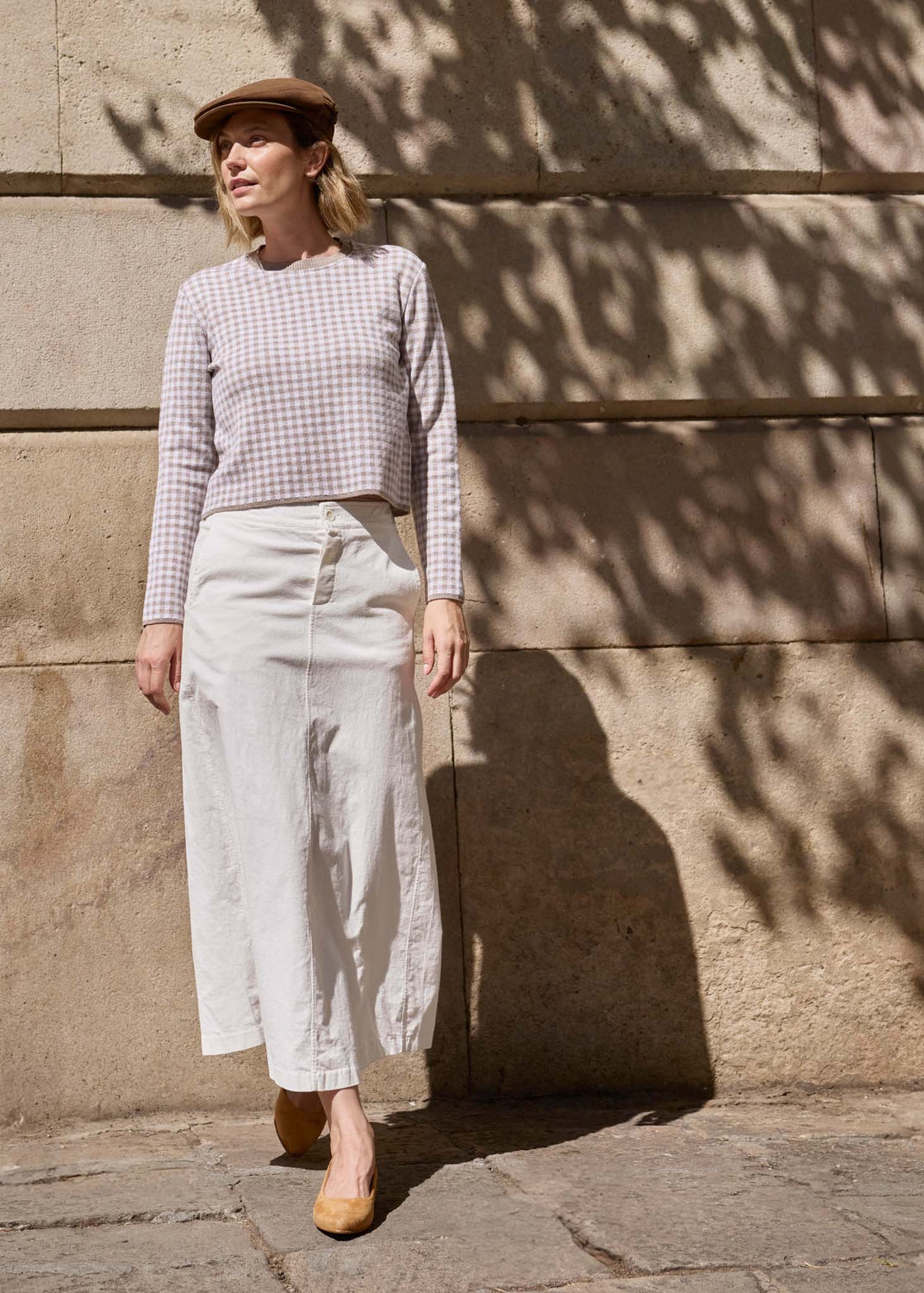 A woman stands outdoors against a stone wall with dappled sunlight and shadows, wearing a beige check long-sleeve top, a cream wide-leg skirt, and tan pointed flats.