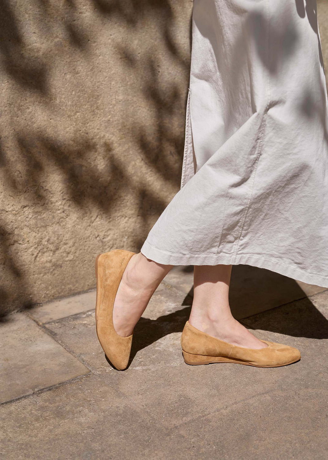 A person wearing light beige wide-leg trousers and tan suede ballet flats, standing on a stone pavement against a textured wall with leaf shadows.