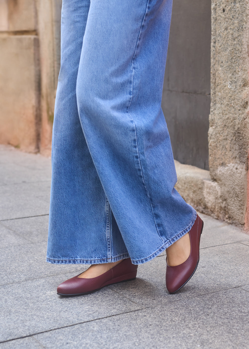 Close-up of woman wearing grain burgundy leather flats with wide-leg blue jeans on a city sidewalk — Viscata Barcelona’s effortless blend of comfort and modern style.