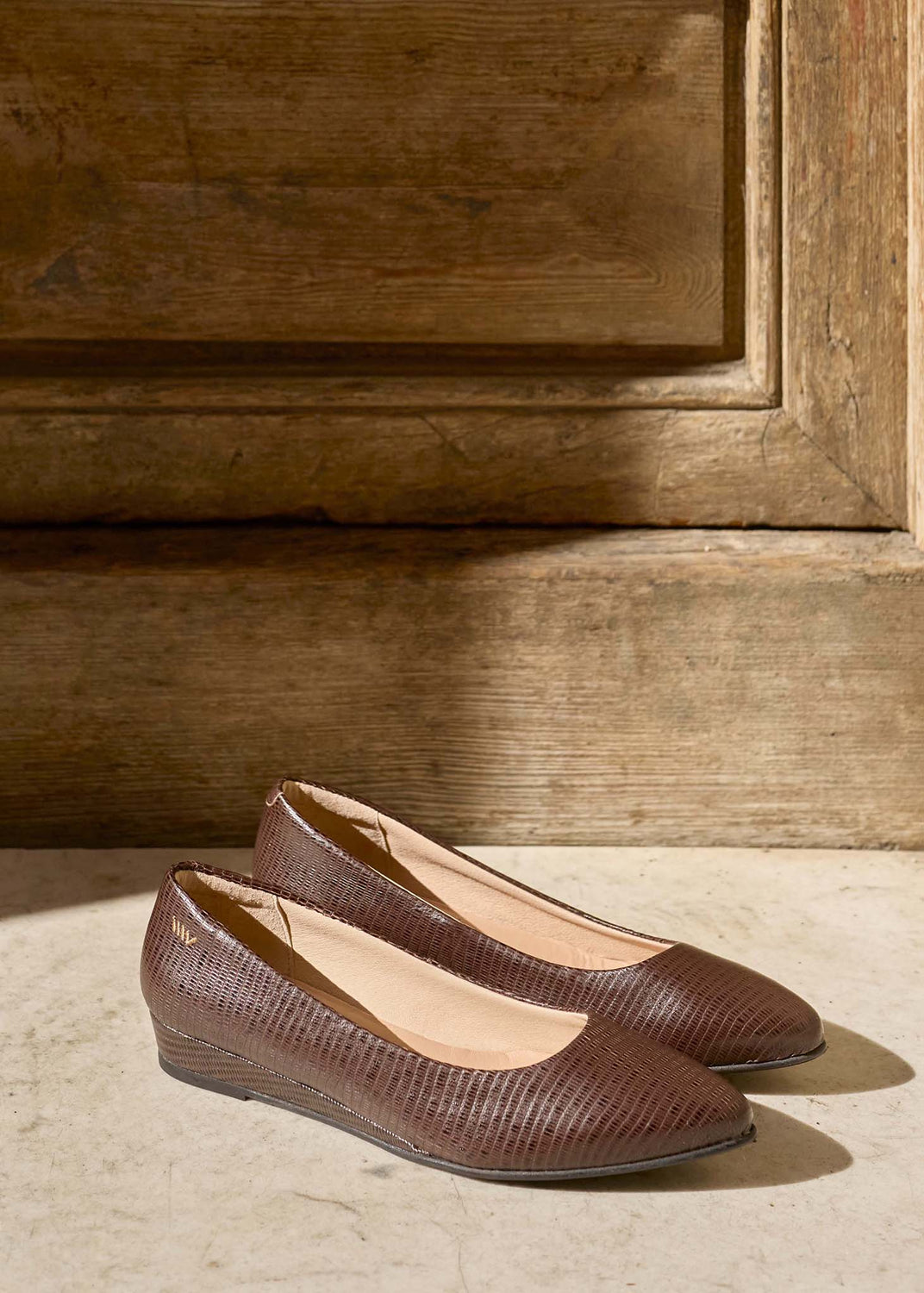 A pair of brown textured wedge flats with pointed toes, displayed on a light stone floor in front of a rustic wooden door.