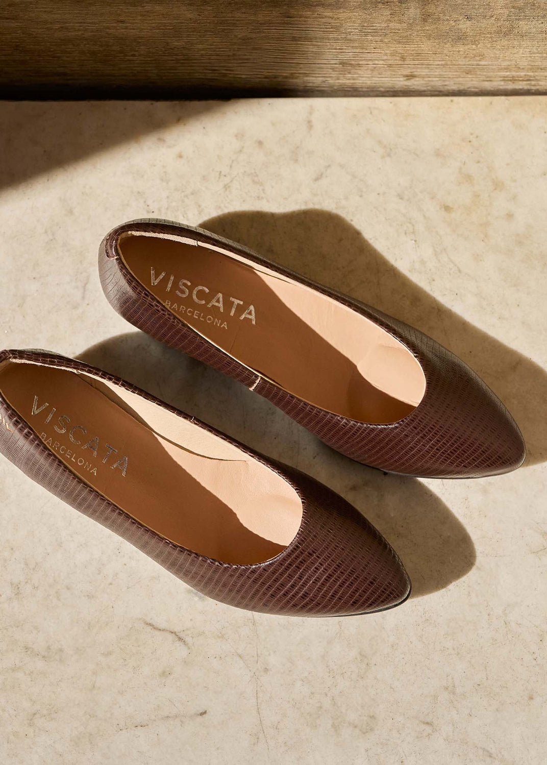 A pair of Viscata brown pointed-toe flats with a textured snakeskin pattern, displayed on a beige stone surface with warm natural shadows.
