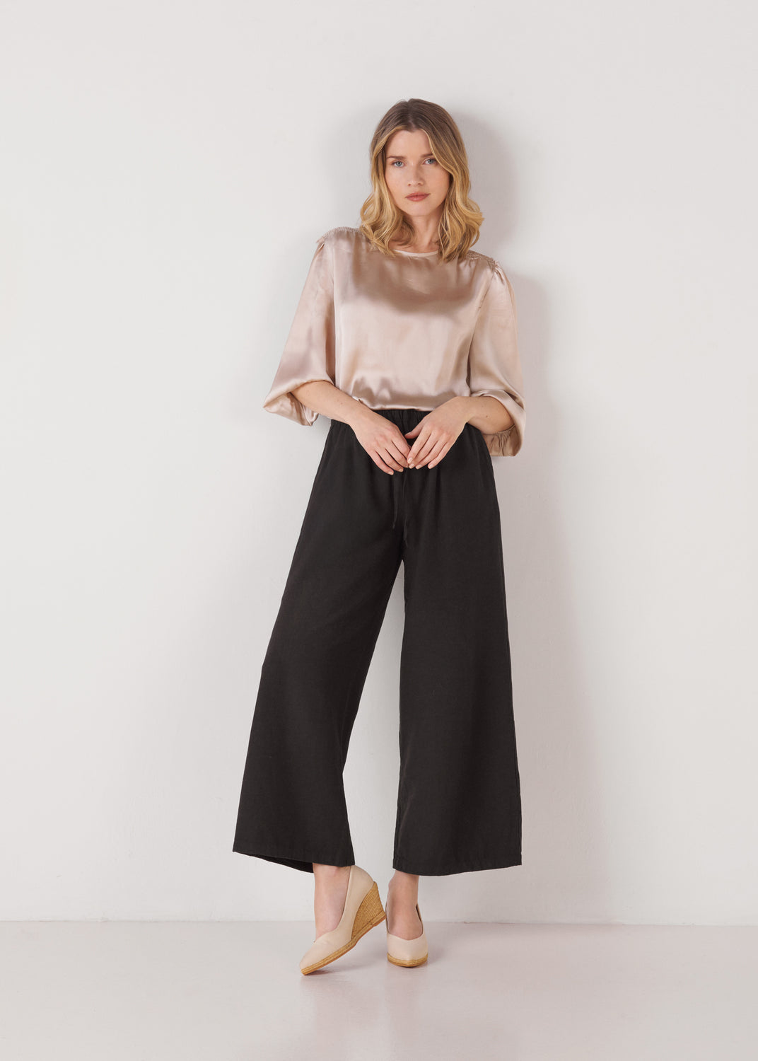 Model standing in beige canvas slip-on wedge espadrilles with a natural jute sole, styled with black wide-leg pants