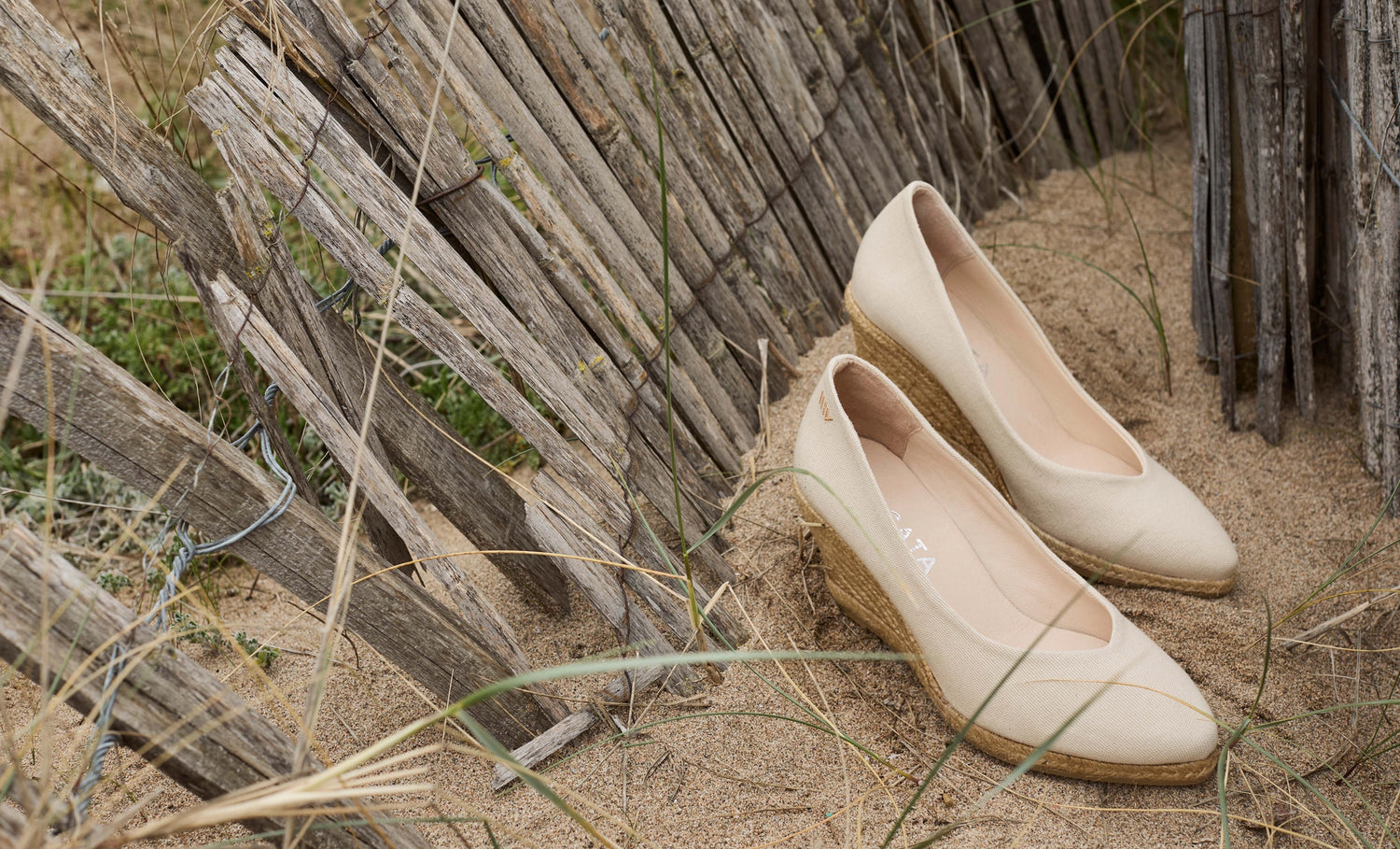 A pair of beige slip-on wedge shoes placed on sand beside a rustic wooden fence.
