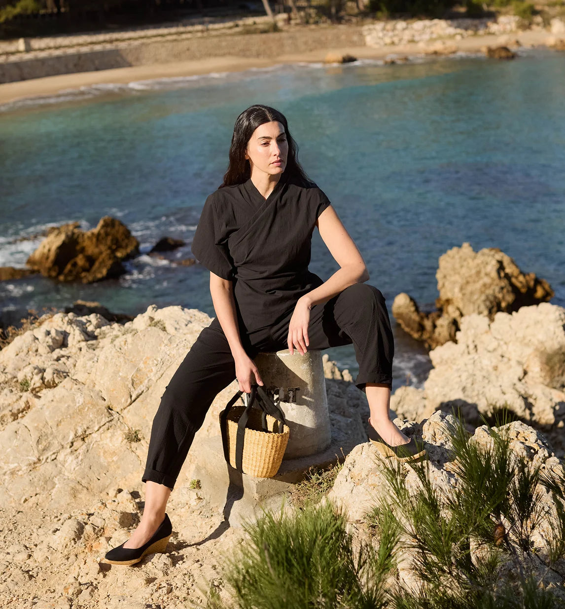 A woman sits on a stone pedestal on a rocky beach, wearing a black short-sleeve wrap-style top and black cropped pants, holding a woven straw handbag.
