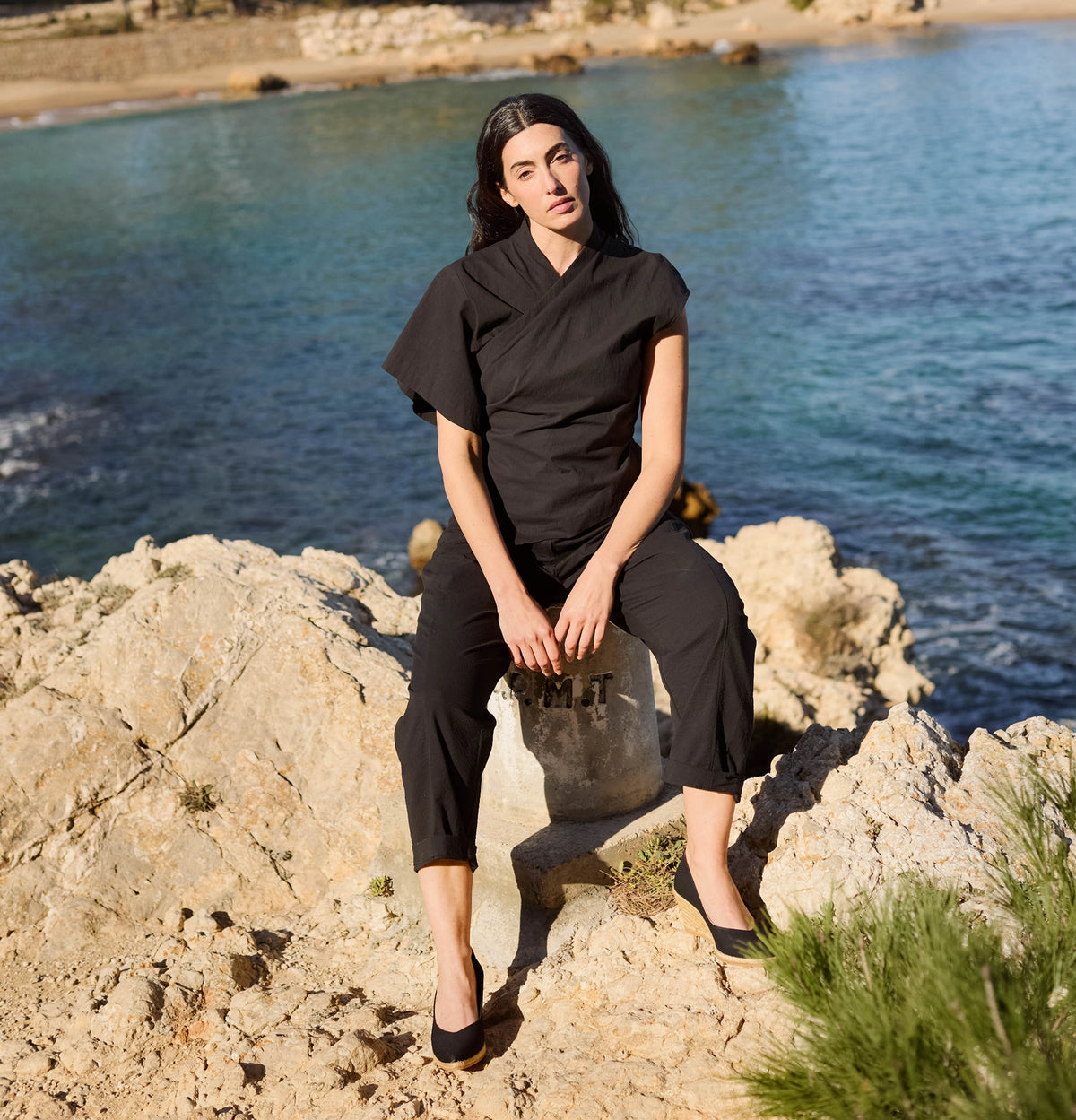 A woman wearing a black short-sleeve wrap-style top and matching cropped pants sits on rocks by the sea, with blue water and shoreline in the background.