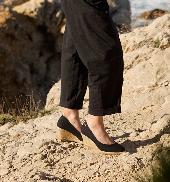 A person wearing black slip-on wedge espadrilles with a woven jute sole, standing on a rocky beach path.