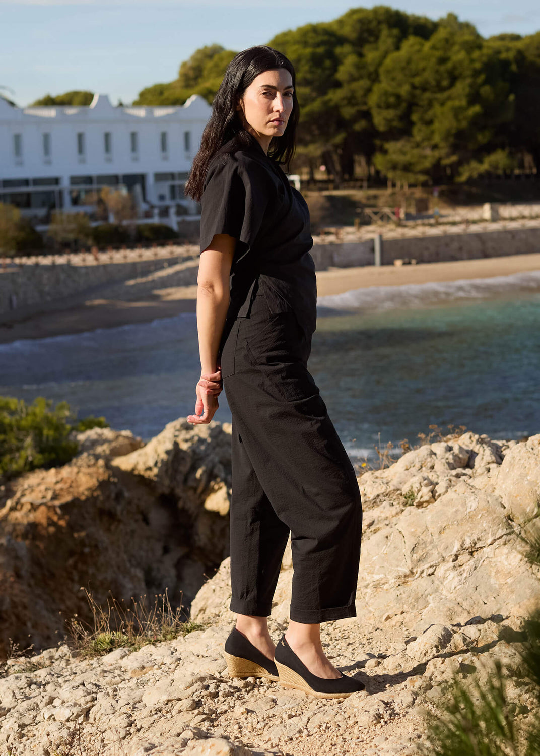 Woman standing on a rocky coastal path overlooking the sea, dressed in an all-black outfit paired with Roses black canvas slip-on wedge espadrilles — a modern expression of Mediterranean simplicity and quiet strength.