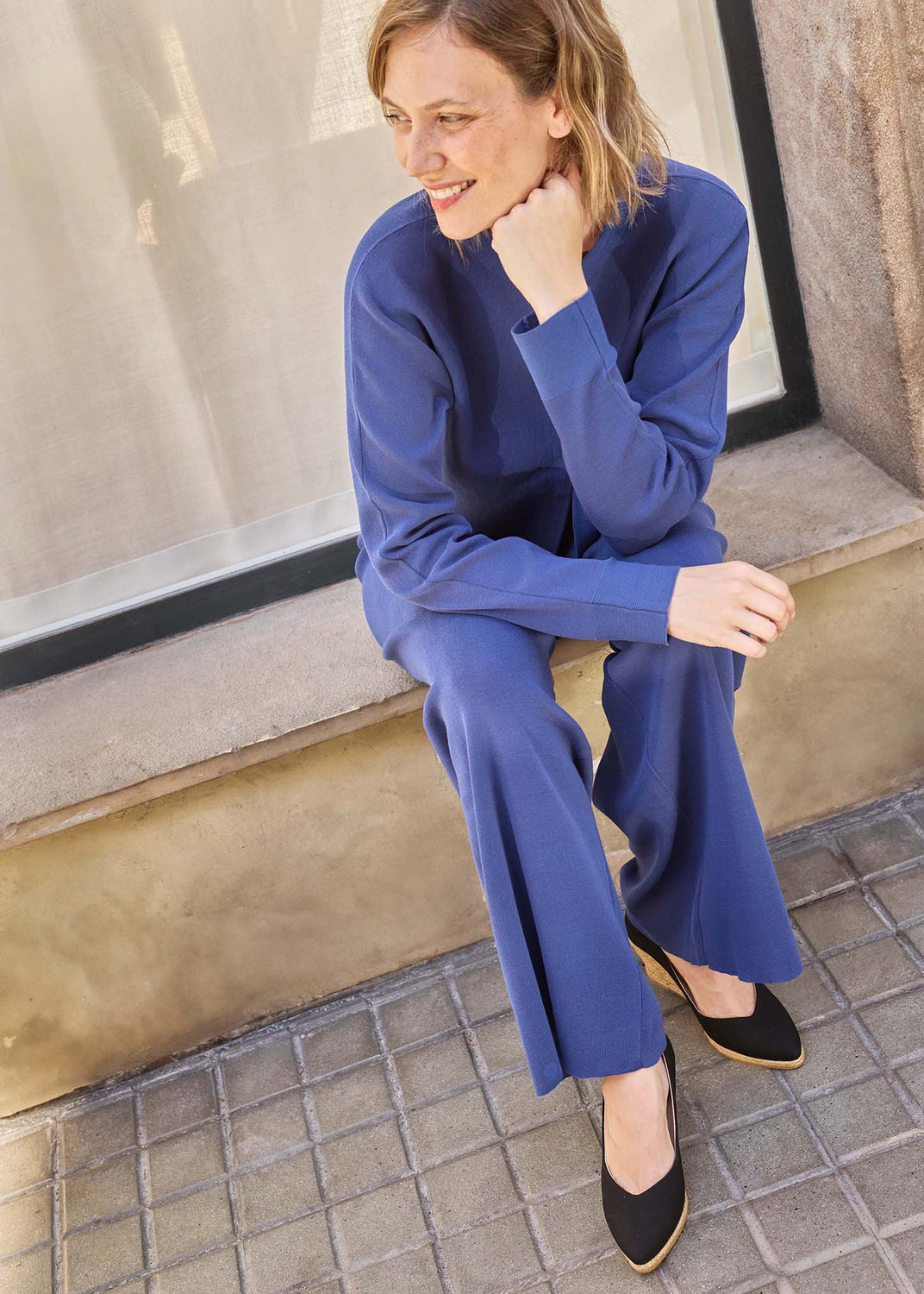 A woman wearing a matching royal blue long-sleeve top and wide-leg trousers sits on a stone ledge next to a window.