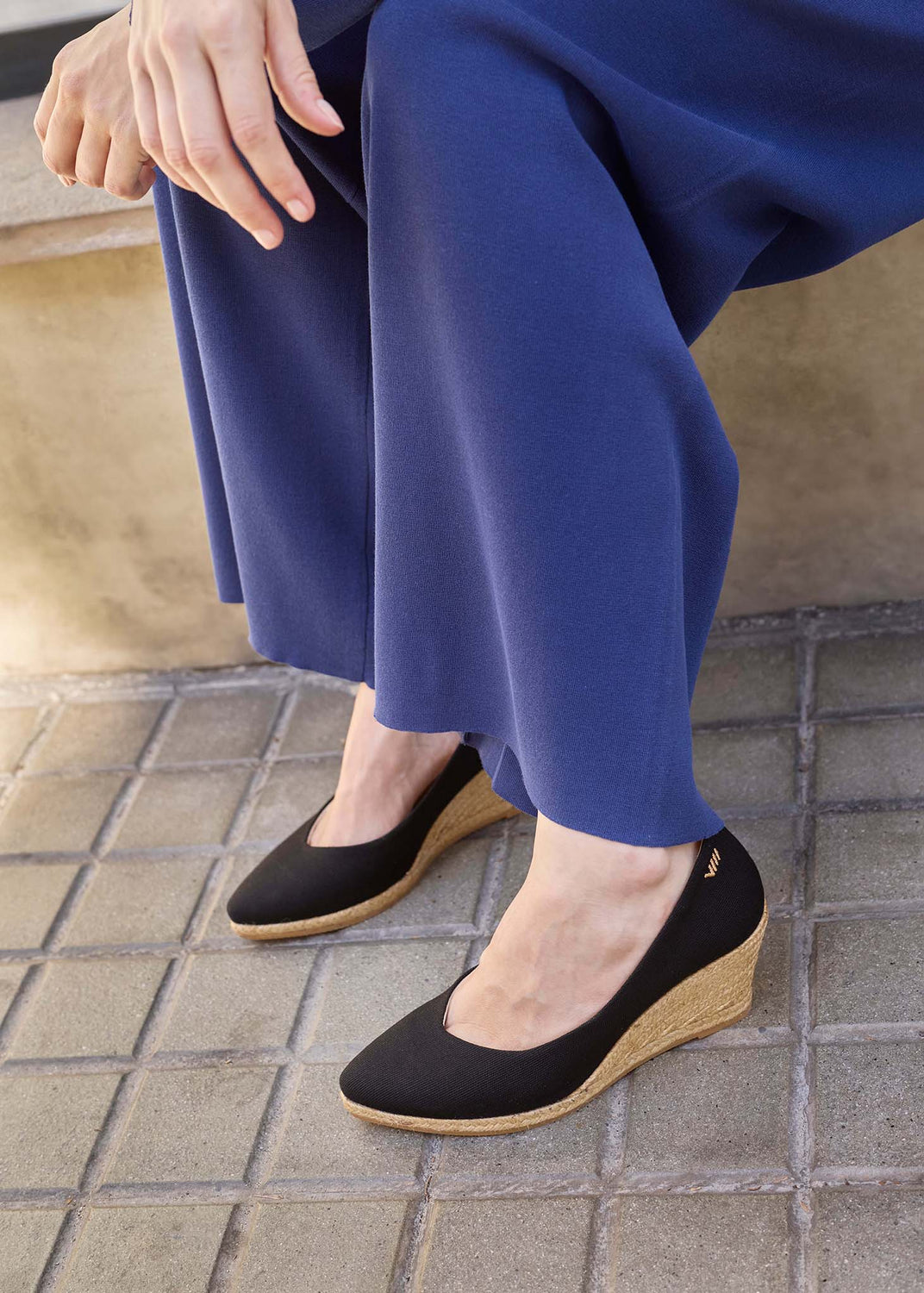 A person seated outdoors wearing black slip-on wedge shoes with a natural jute-wrapped sole and blue wide-leg pants, photographed from the knees down on a stone-tiled surface.