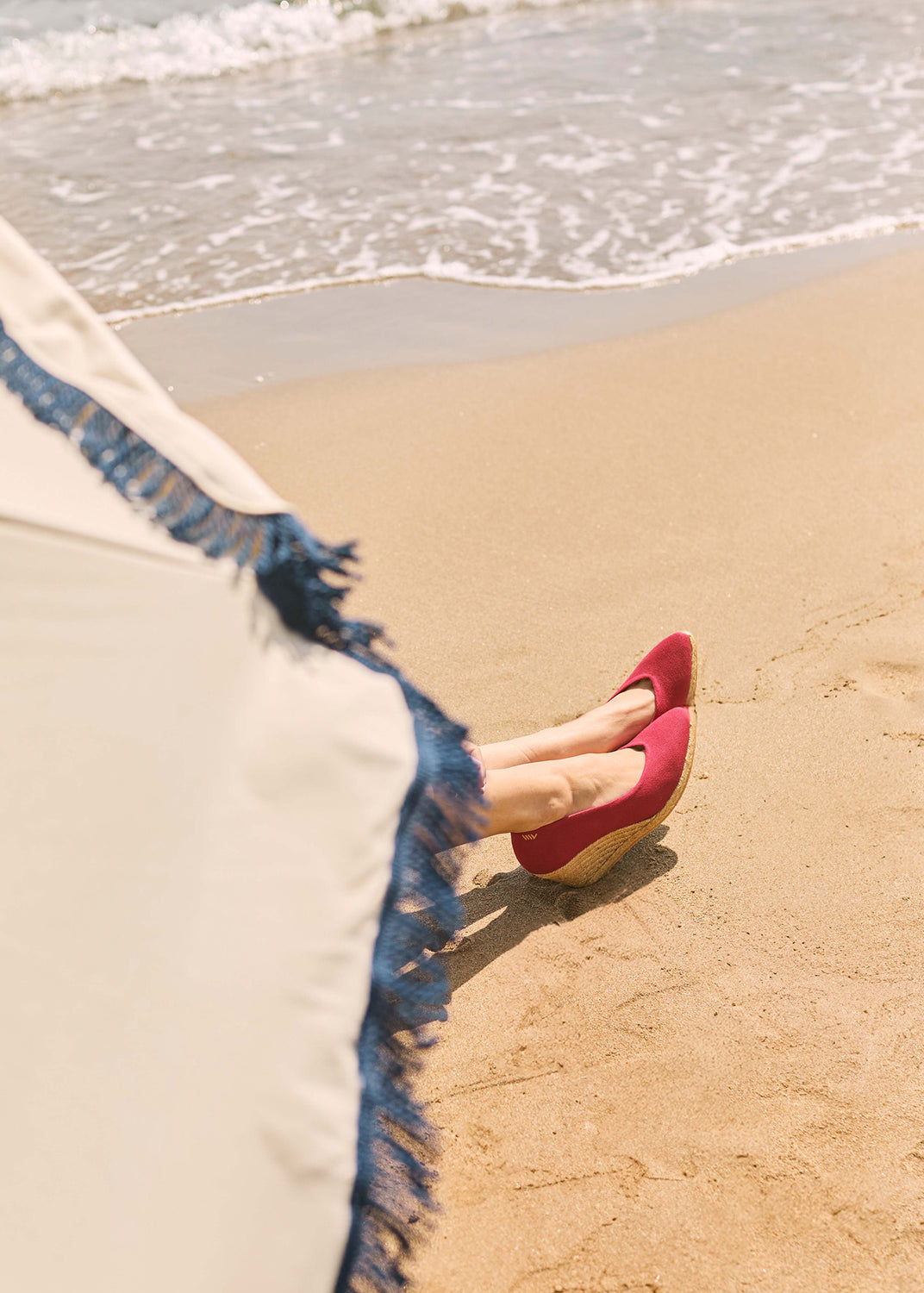 Cherry red wedge espadrilles resting on the sand beside the shoreline with a fringed beach umbrella in a Mediterranean coastal setting.