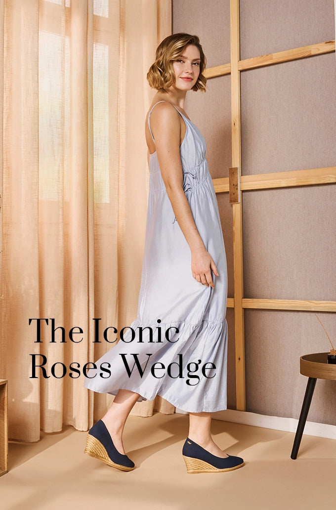Woman in a light blue maxi dress wearing Viscata’s iconic Roses wedge espadrilles in navy canvas with natural jute heels, standing indoors beside beige curtains and wooden decor.