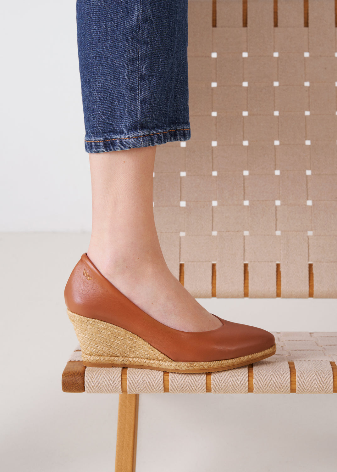 Close-up of whiskey leather slip-on wedge espadrille on model, with natural jute sole, resting on a woven chair