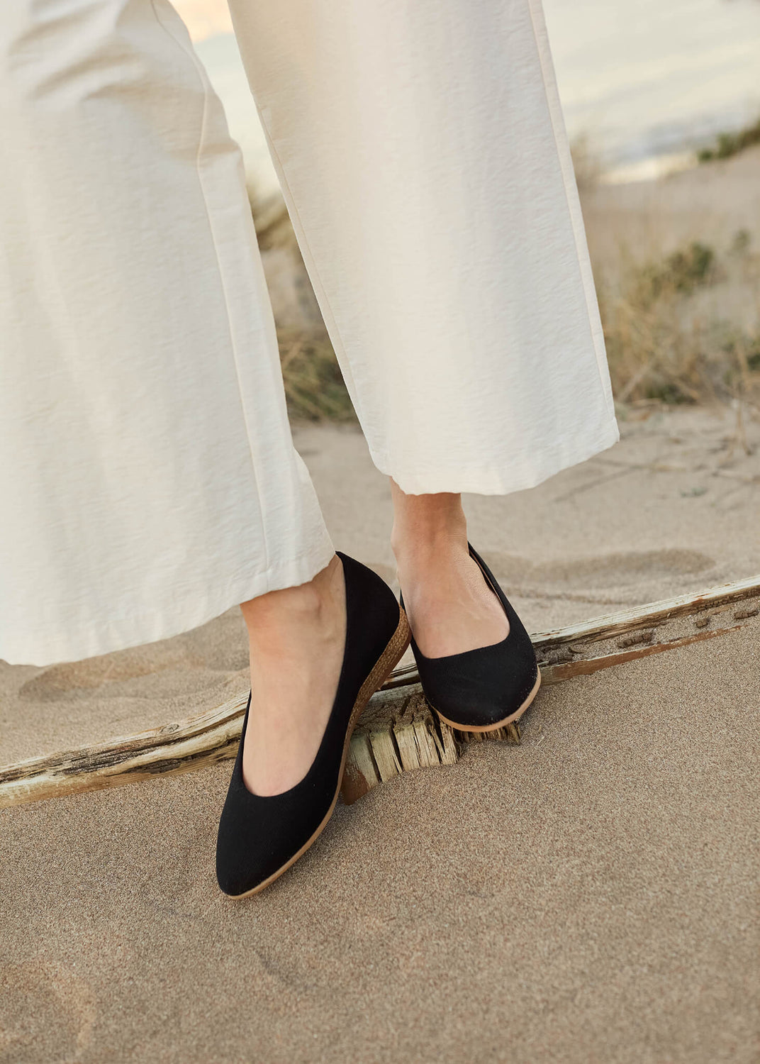 Close-up of a woman standing barefoot on soft coastal sand, wearing wide-leg ivory trousers and Rubina black canvas slip-on flats — an effortless expression of timeless Mediterranean ease.
