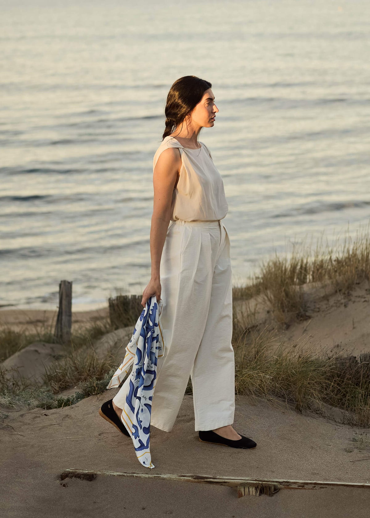 Woman standing by the shore at sunset wearing Rubina black canvas slip-on flats, cream trousers, and holding a blue-and-white silk scarf, evoking effortless coastal elegance.