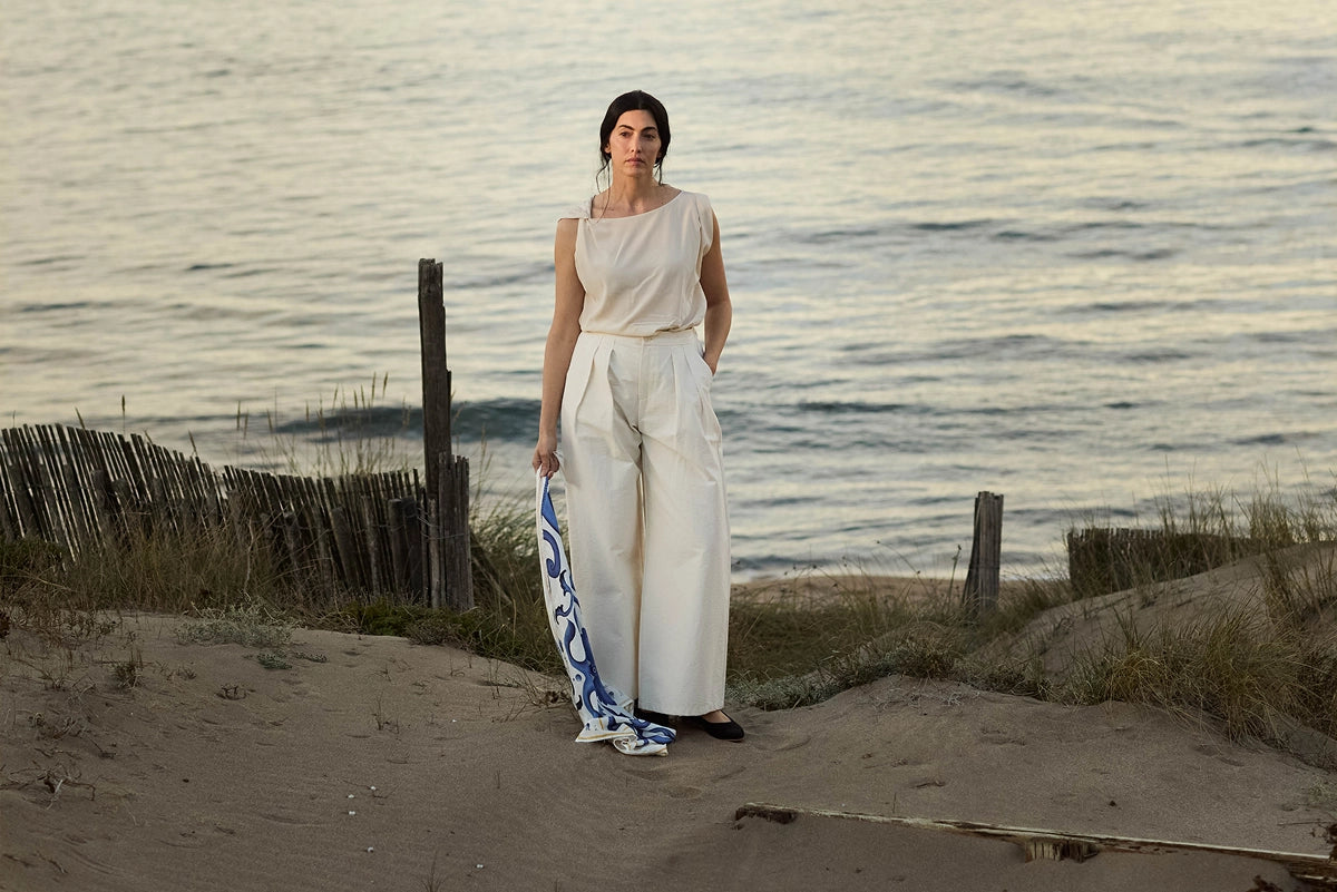 A woman stands on a sandy dune by the sea, wearing a sleeveless cream top and matching wide-leg trousers, holding a blue and white patterned scarf.