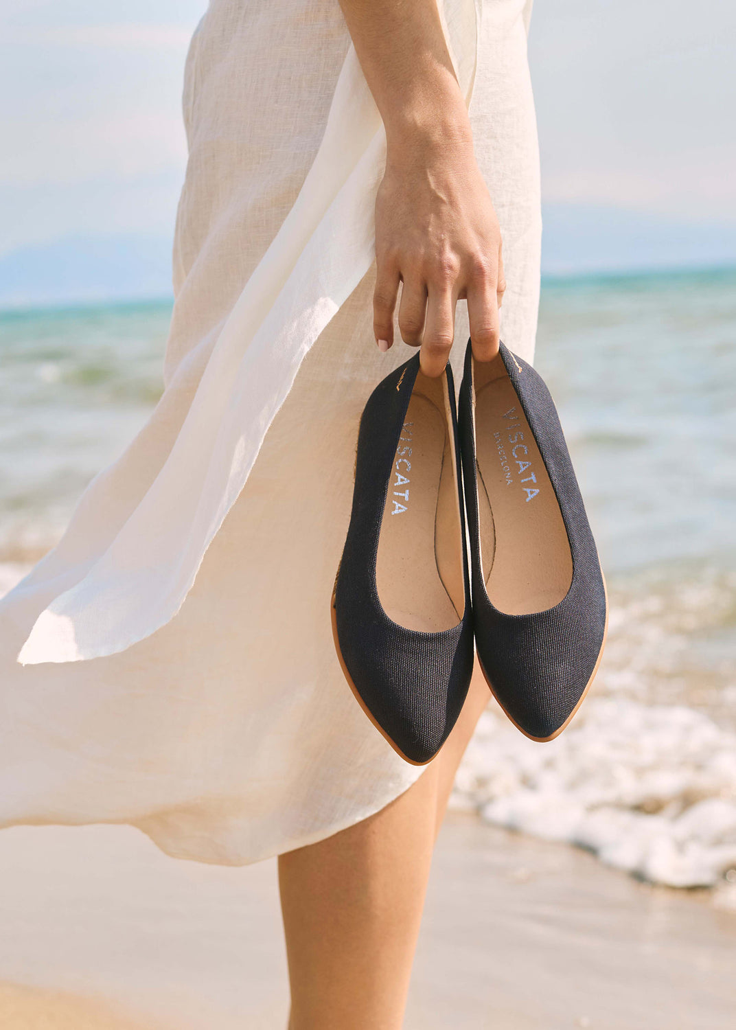 Navy blue espadrille flats held by hand beside the shoreline in a Mediterranean beach setting.