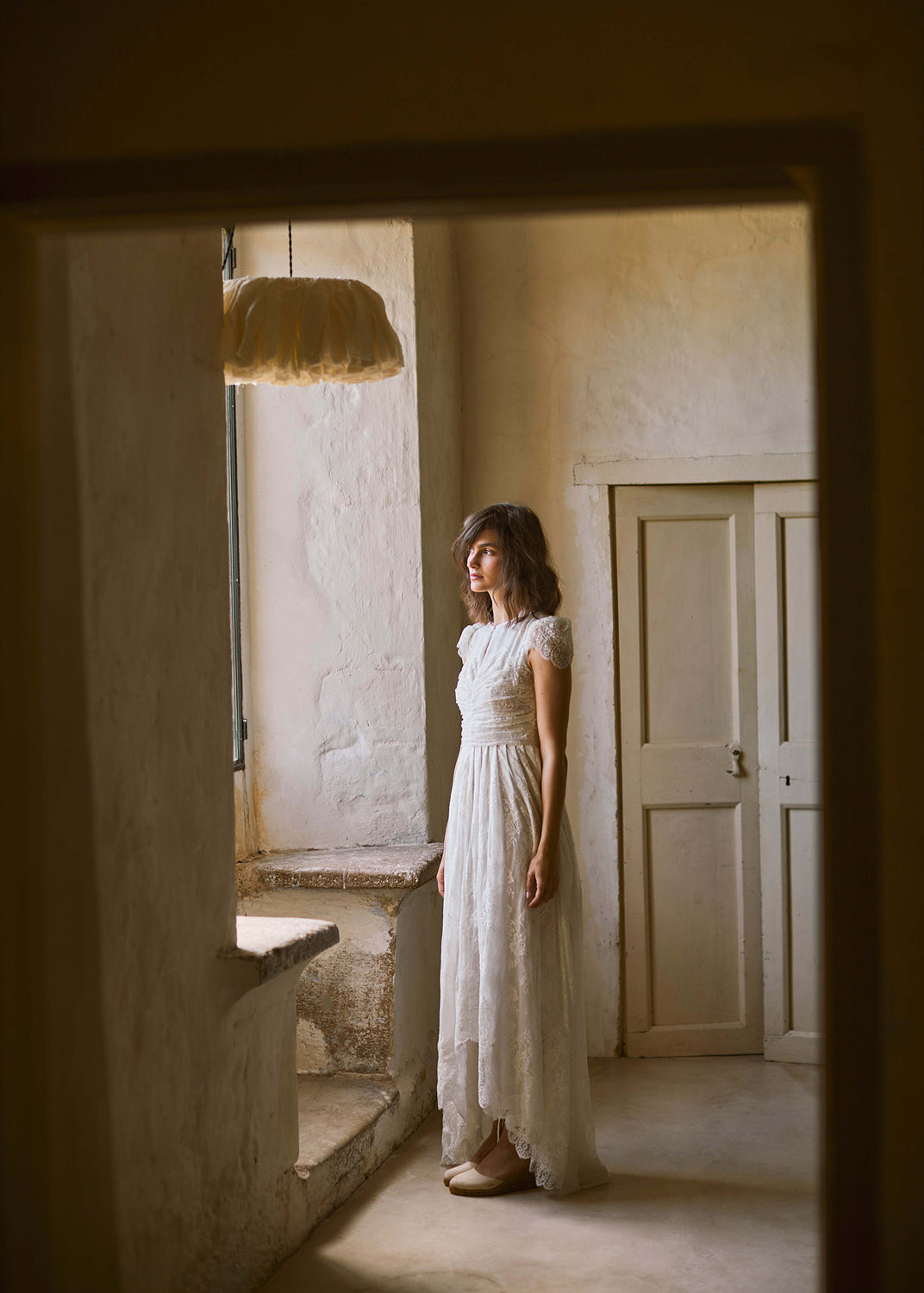 Woman in a white lace dress wearing beige espadrille wedges in a softly lit Mediterranean interior.