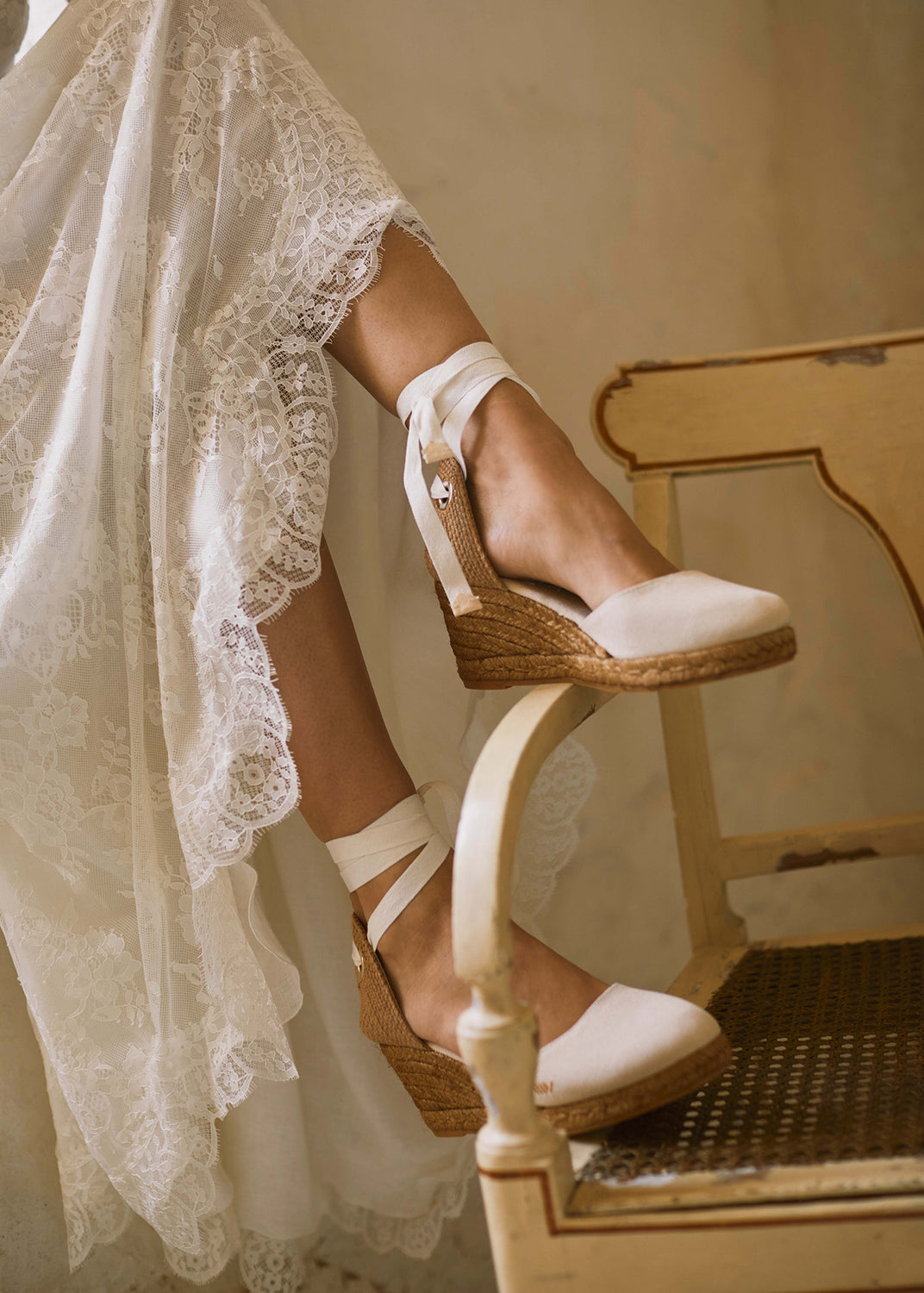 Beige lace-up wedge espadrilles styled beneath a flowing lace dress in a warm Mediterranean interior.