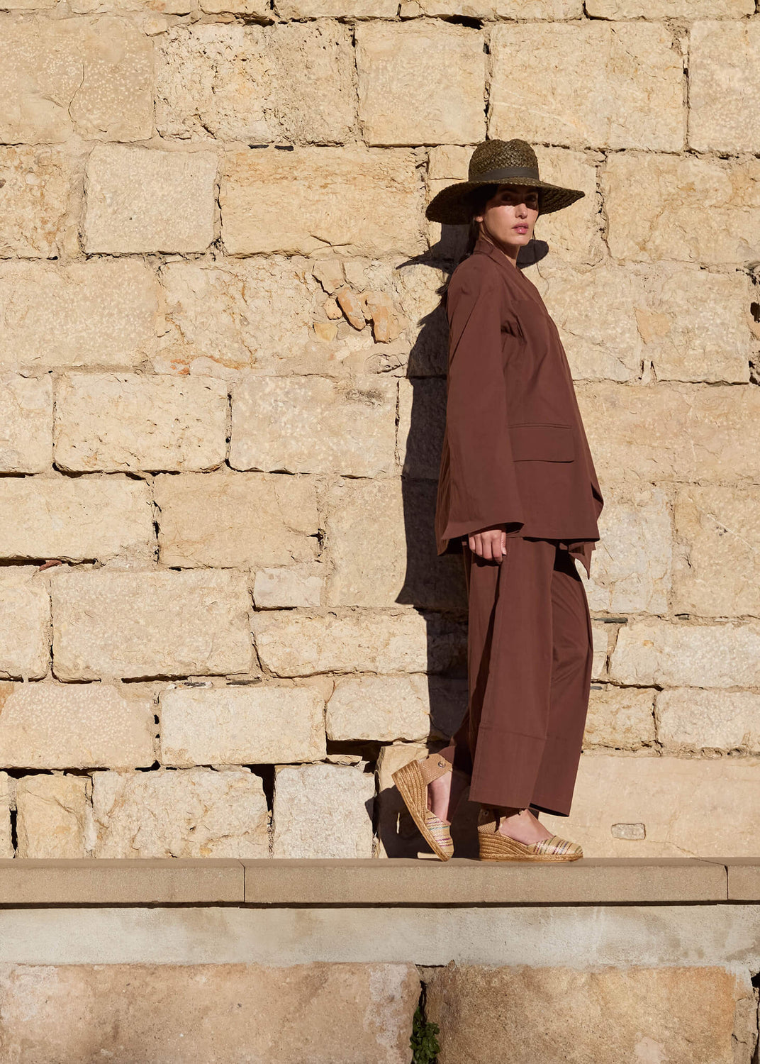Woman standing against a sunlit stone wall, dressed in an earthy brown ensemble and wearing Sagaro multicolor raffia lace-up wedge espadrilles. The soft light and natural textures evoke a grounded, Mediterranean ease.