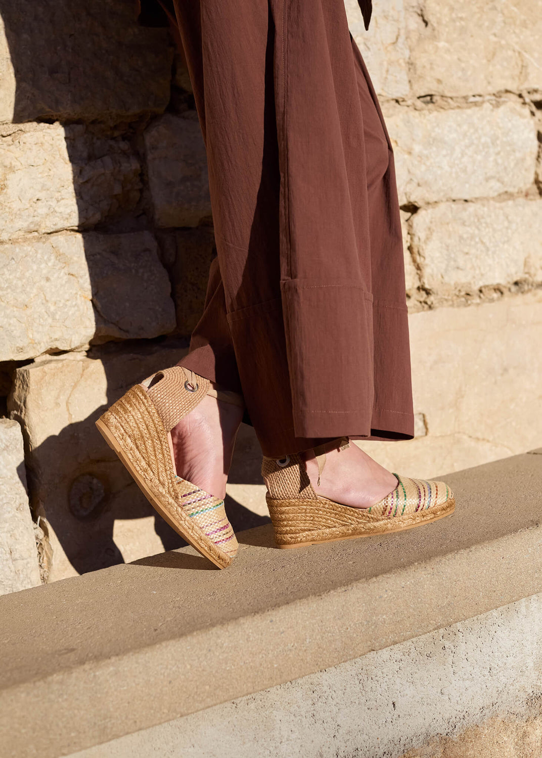Close-up of a woman standing by a sunlit stone wall, wearing Sagaro multicolor raffia lace-up wedge espadrilles paired with flowing brown trousers, capturing warm textures and effortless Mediterranean charm.