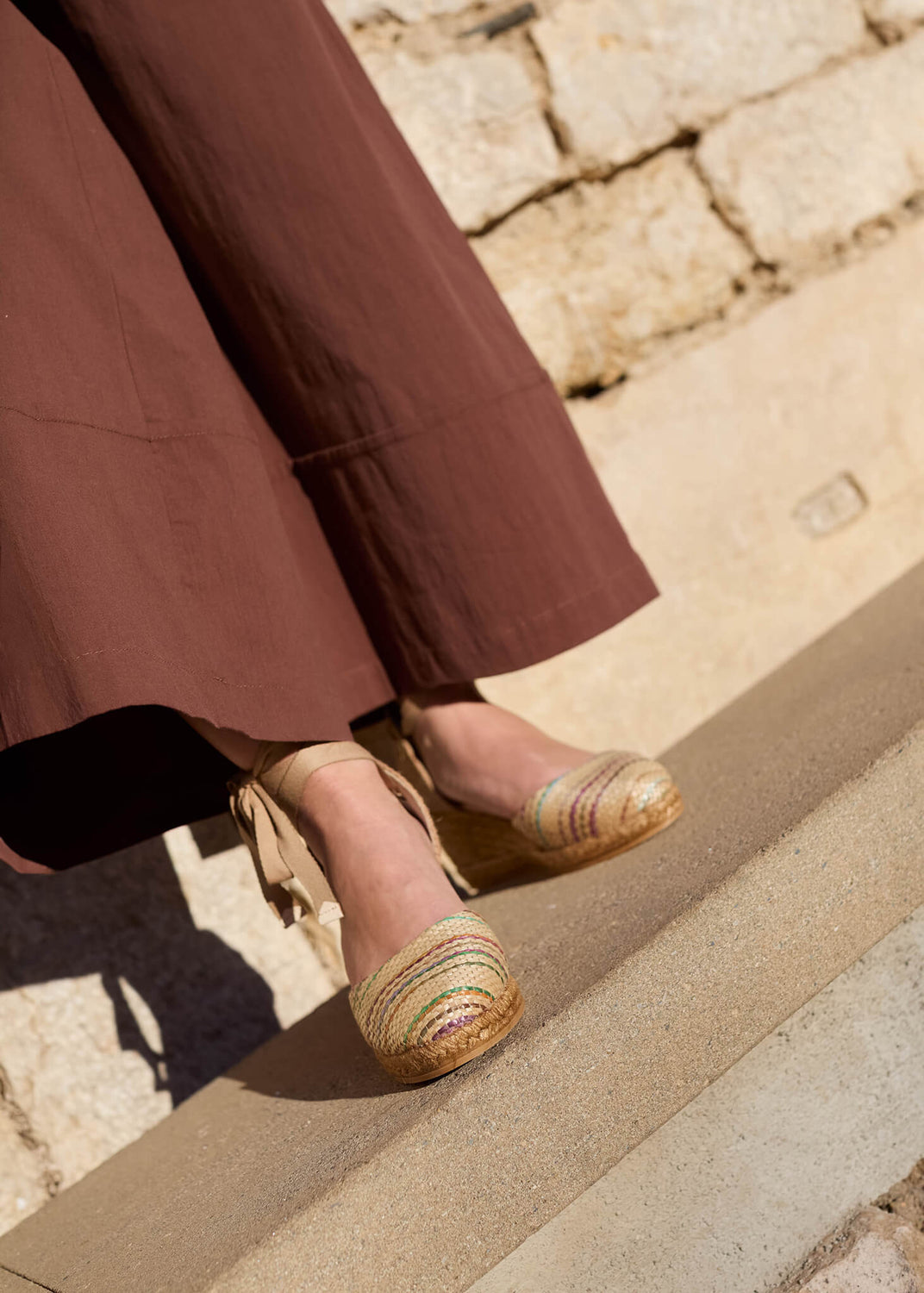 Close-up of a woman wearing Sagaro multicolor raffia lace-up wedge espadrilles, paired with wide brown trousers under warm Mediterranean light.