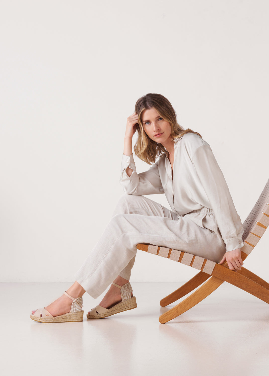 Woman sitting on a wooden lounge chair wearing beige canvas ankle-strap wedge espadrille sandals in a bright, minimalist studio.