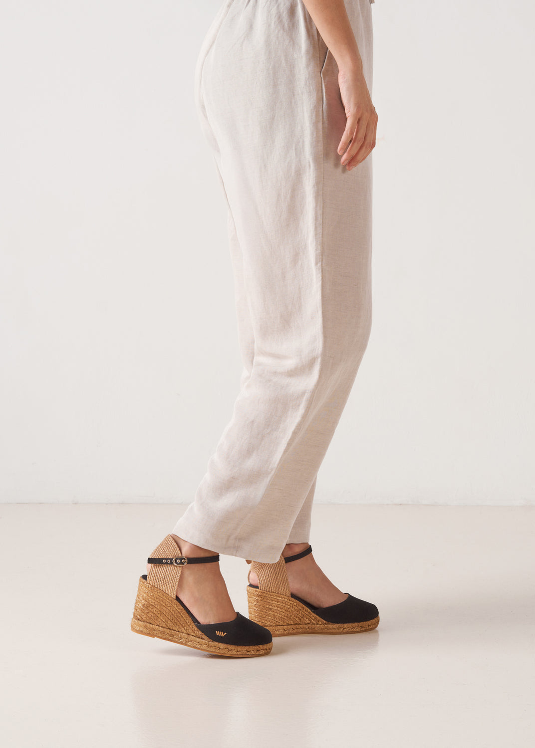 Side view of a woman wearing black canvas ankle-strap wedge espadrilles with jute soles in a clean, white studio.