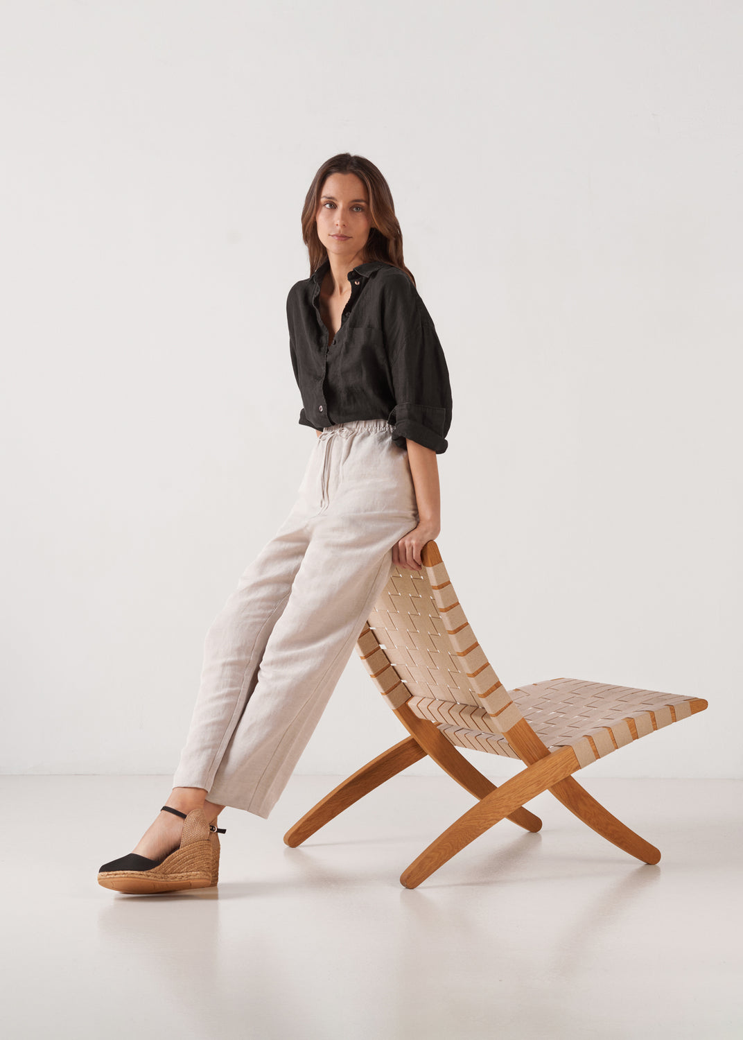 Full-body studio shot of model leaning on chair wearing black ankle-strap wedge espadrilles.