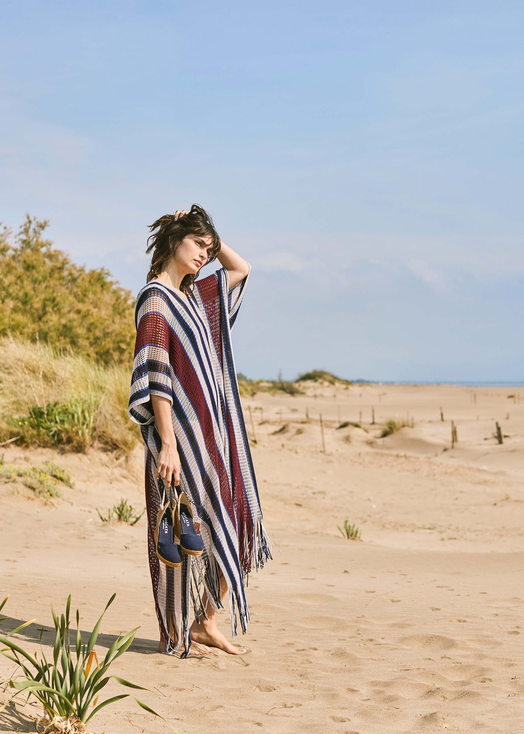 Woman standing on Mediterranean sand dunes wearing a striped kaftan and holding navy blue canvas espadrille wedges with ankle straps.