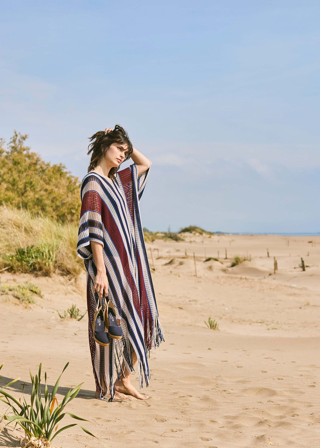 Woman standing on Mediterranean sand dunes wearing a striped kaftan and holding navy blue canvas espadrille wedges with ankle straps.