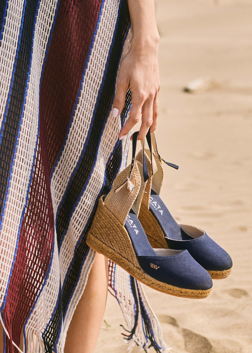 Navy blue canvas espadrille wedges with slingback straps held by hand on Mediterranean sand dunes beside a striped beach kaftan.