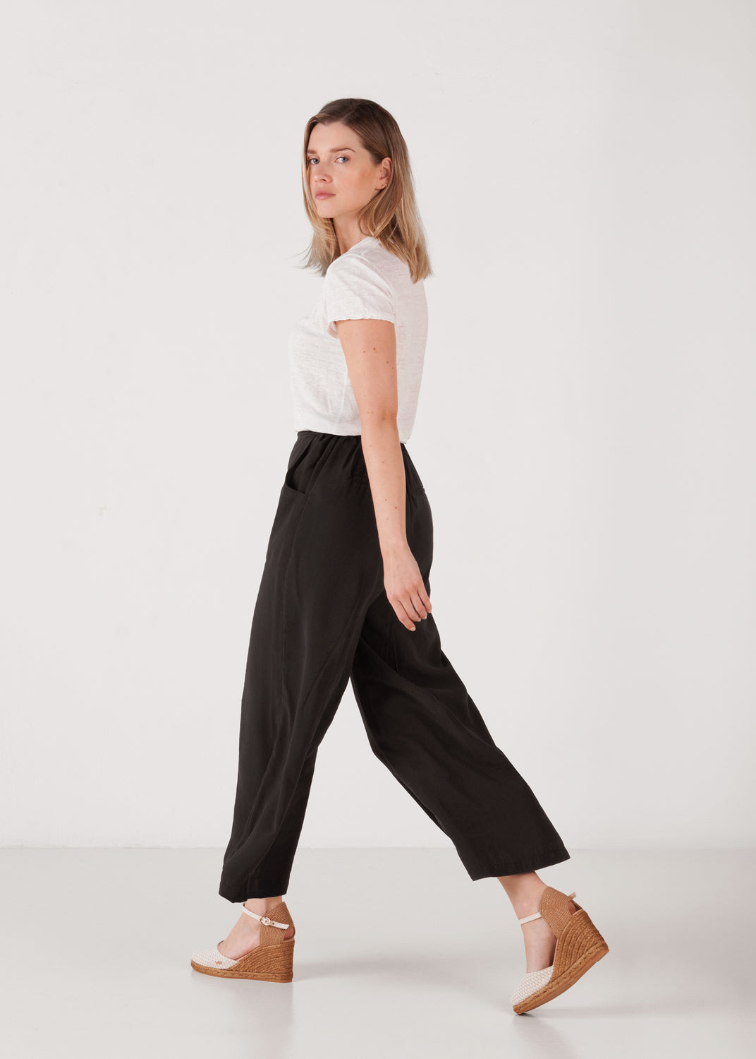 Model walking in white mesh-canvas ankle-strap wedge espadrilles for women, with jute wedge sole, styled with a white tee and black pants against a minimal studio background.