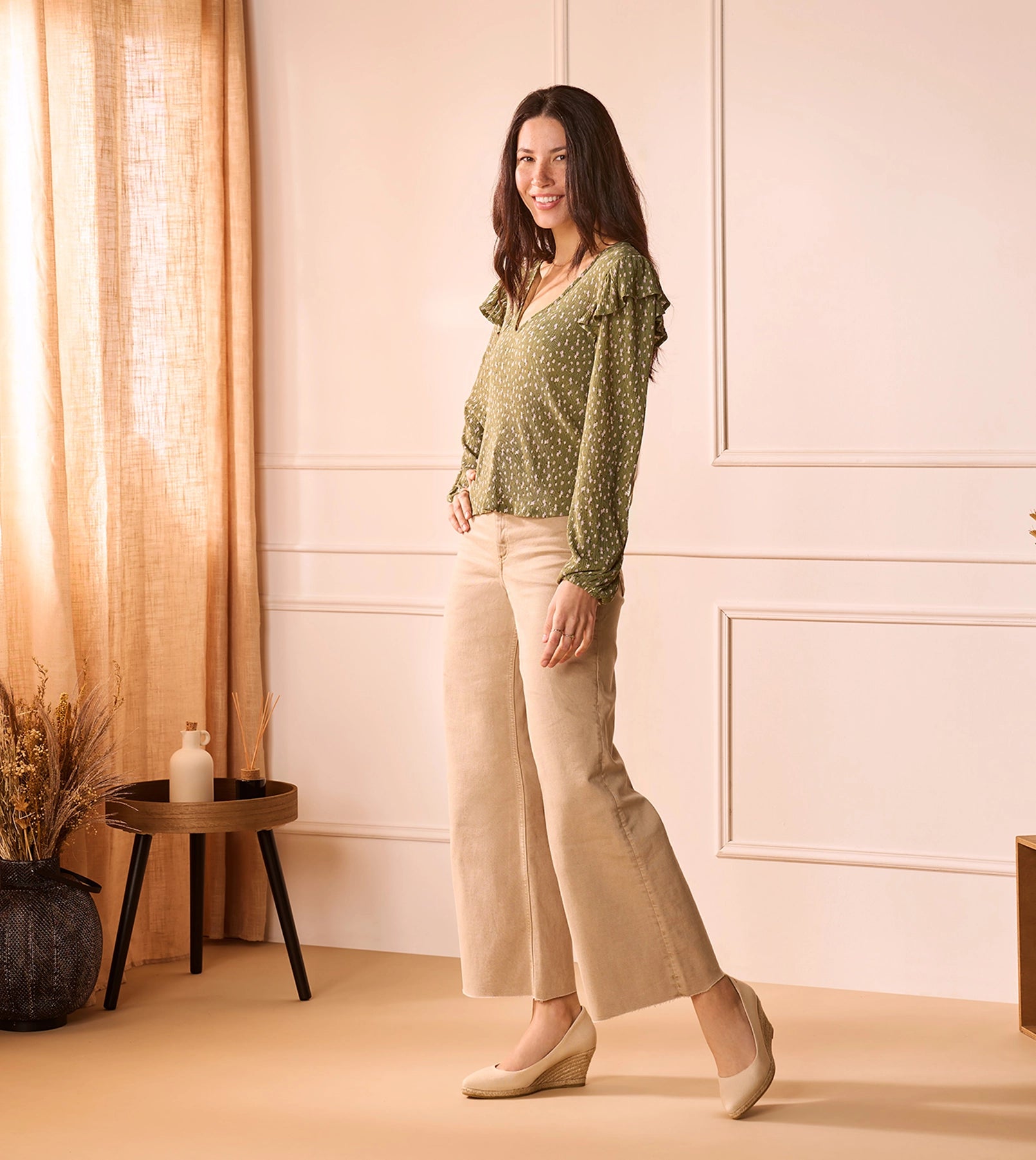 Woman smiling while modeling Viscata beige wedge espadrilles, paired with wide-leg beige pants and a green patterned blouse, standing in a softly lit room with neutral decor.