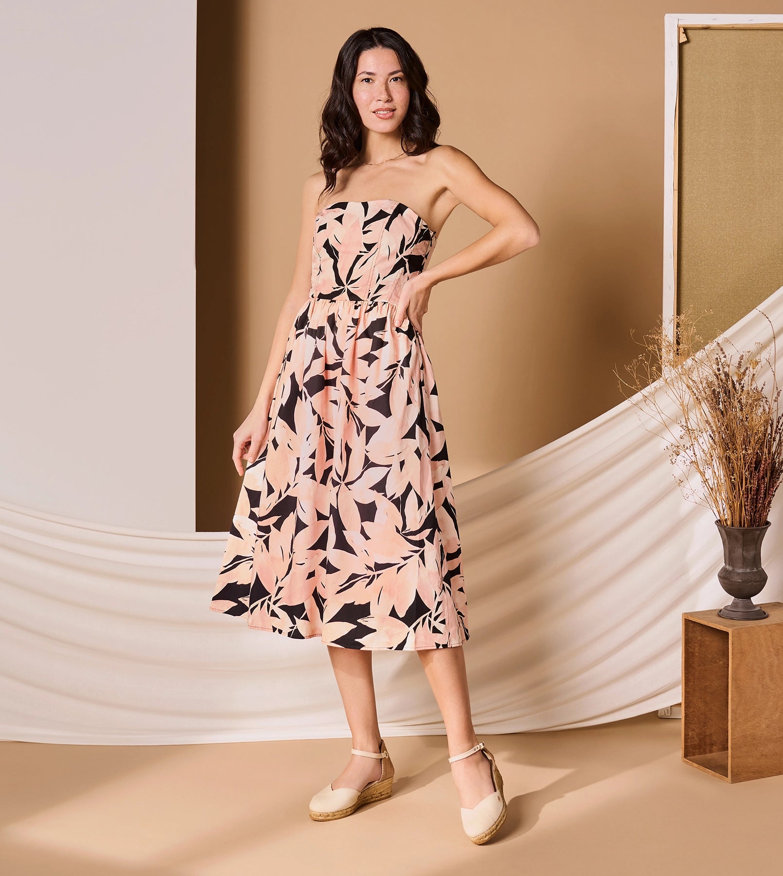 Woman wearing a strapless, knee-length dress with a pink, peach, and black abstract leaf pattern. She is standing in a styled studio with beige and white draped fabric, a neutral backdrop, and dried floral arrangements. She is wearing espadrille wedges with ankle straps and smiling confidently