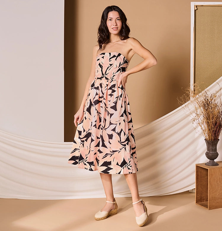 Woman wearing a strapless, knee-length dress with a pink, peach, and black abstract leaf pattern. She is standing in a styled studio with beige and white draped fabric, a neutral backdrop, and dried floral arrangements. She is wearing beige espadrille low wedges with ankle straps and smiling confidently