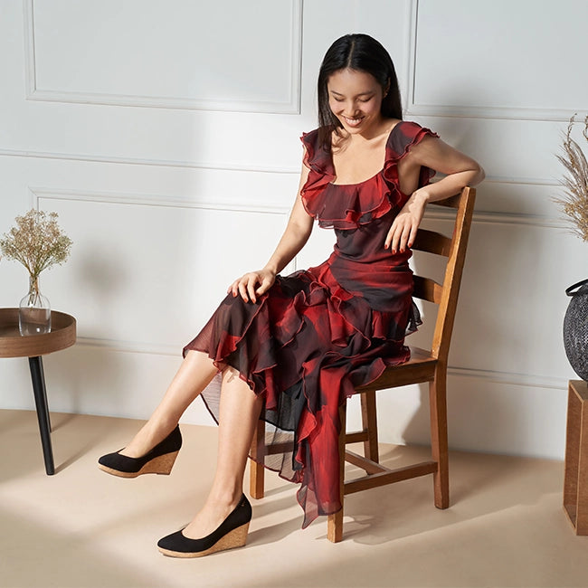 A woman with long dark hair sits on a wooden chair, smiling while looking down. She wears a flowing red and black ruffled dress with sheer fabric and a layered design. Her legs are crossed at the ankles, showcasing black wedge shoes with a cork heel