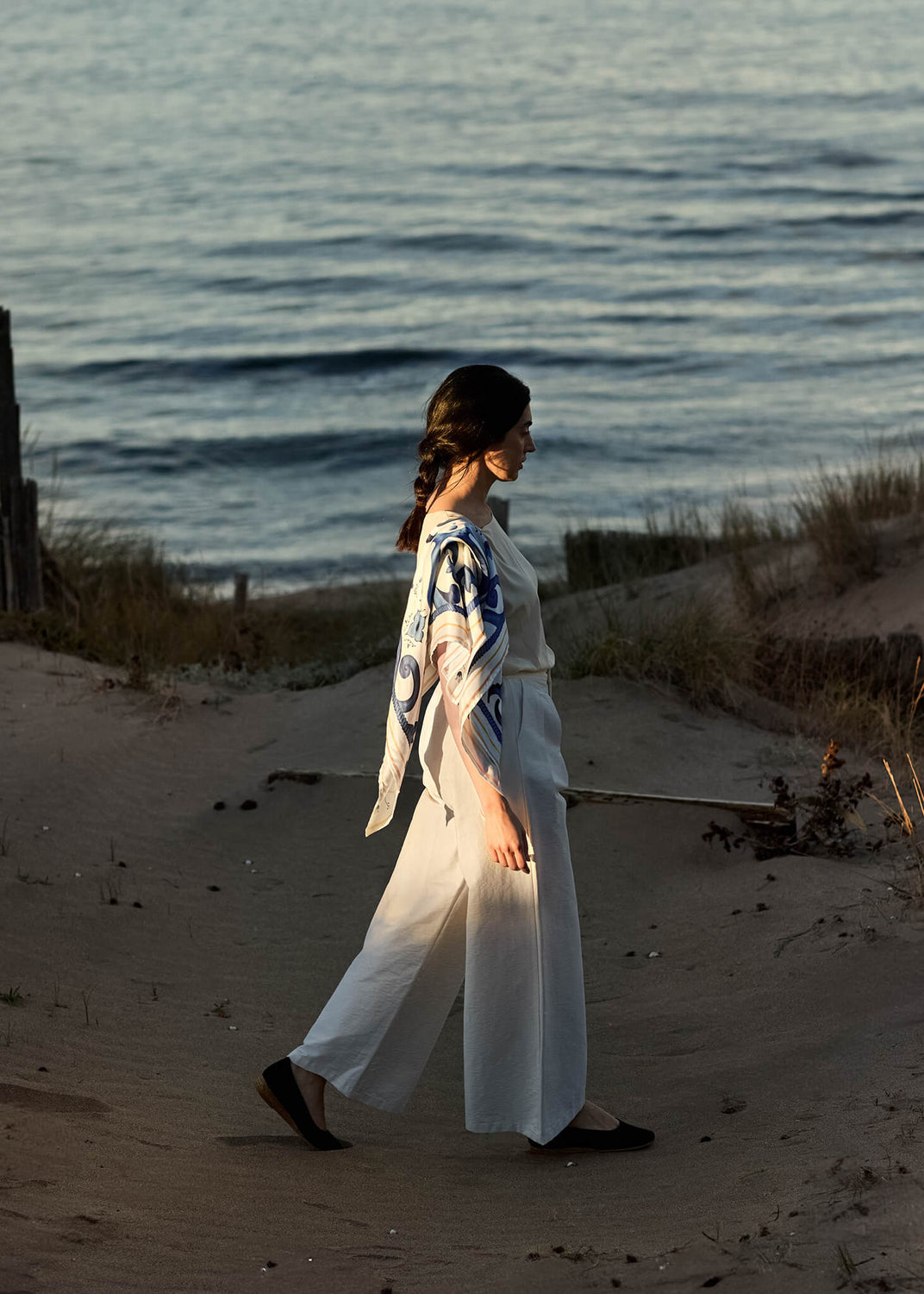 Woman walking barefoot on a sandy path by the sea at sunset, wearing light linen trousers and a cream blouse, with a Majolica Blue square scarf draped elegantly over her shoulders.