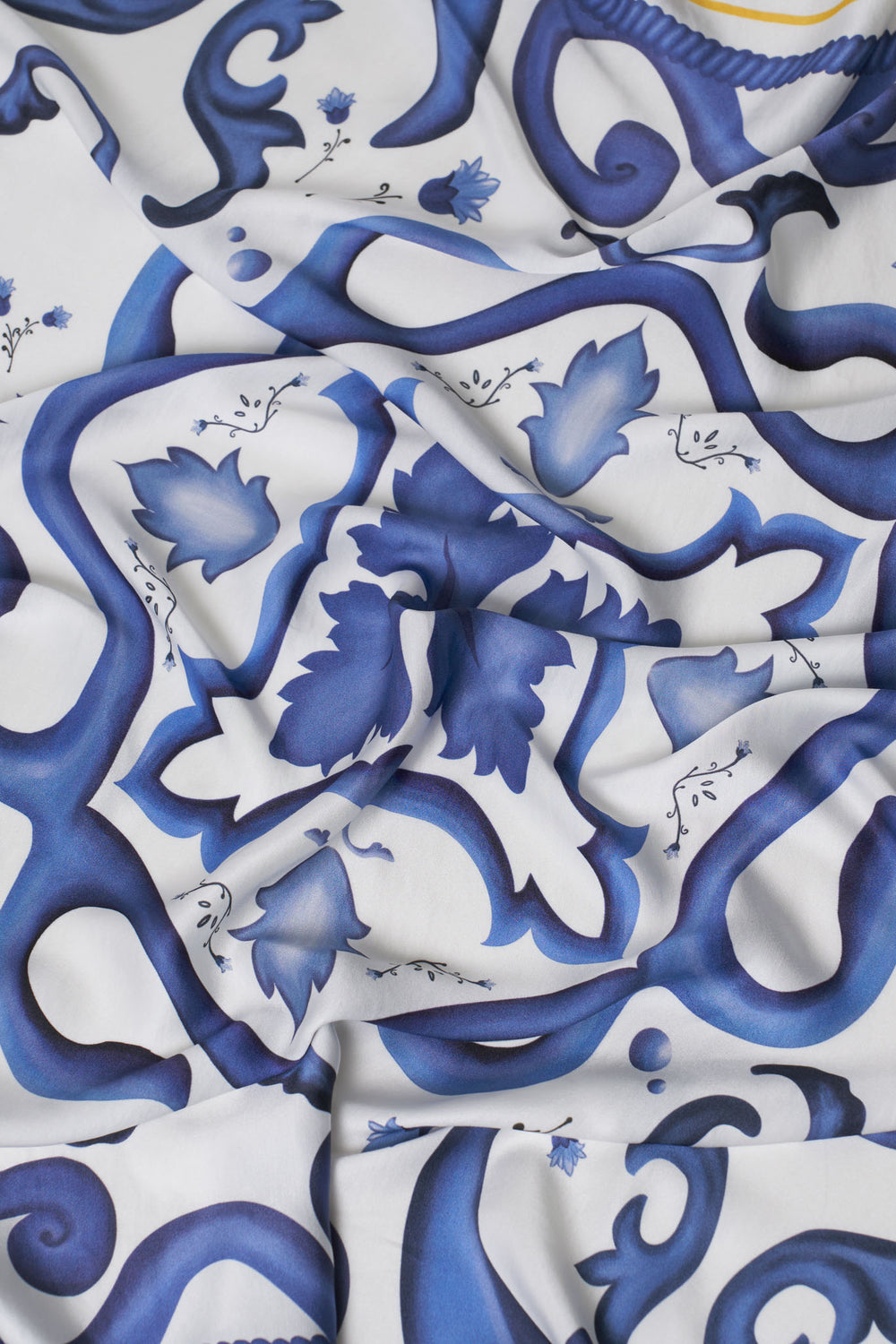 Close-up of flowing carré scarf with deep blue baroque-style floral and scroll design on white background – elegant Mediterranean-inspired accessory highlighting smooth texture and artistic detail