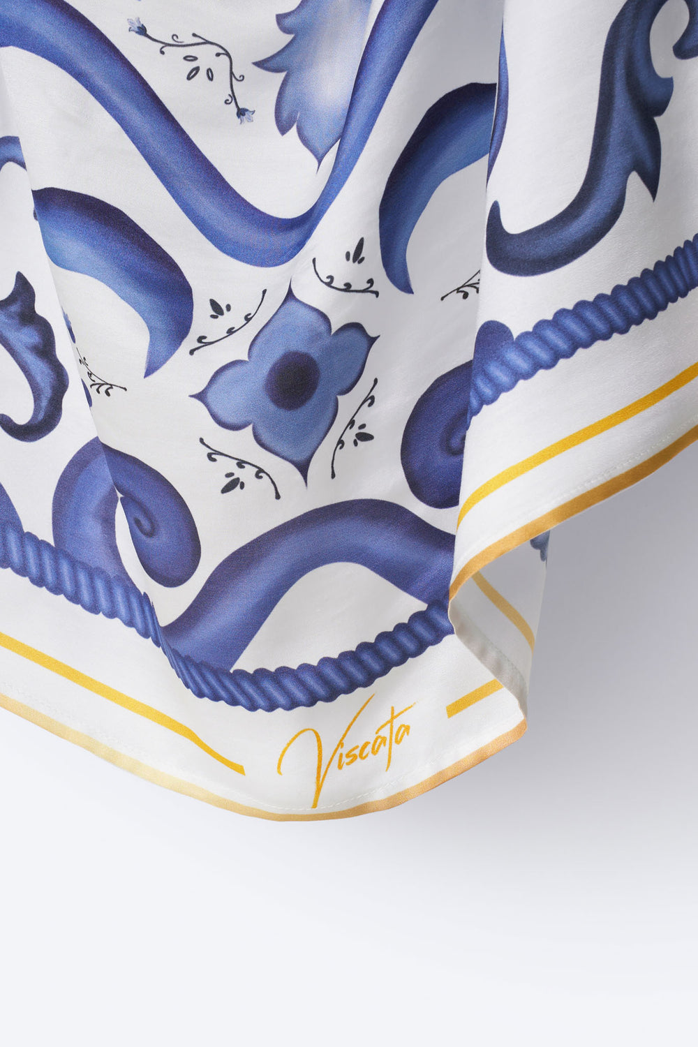 Detail view of Viscata silk scarf corner showcasing signature branding, golden yellow trim, and ornate cobalt blue floral tile pattern – refined Mediterranean accessory with artisan finish