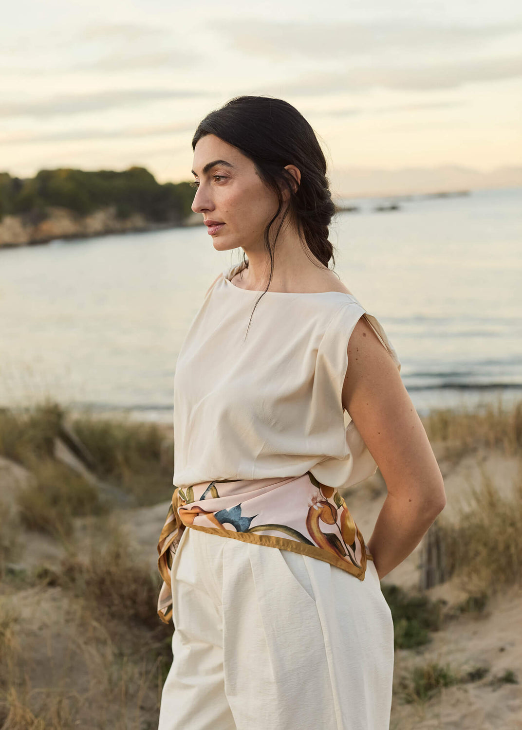 Woman standing by the coast at sunset wearing an ivory sleeveless blouse and cream trousers, styled with a silk scarf in soft caramel and pastel tones tied around her waist.