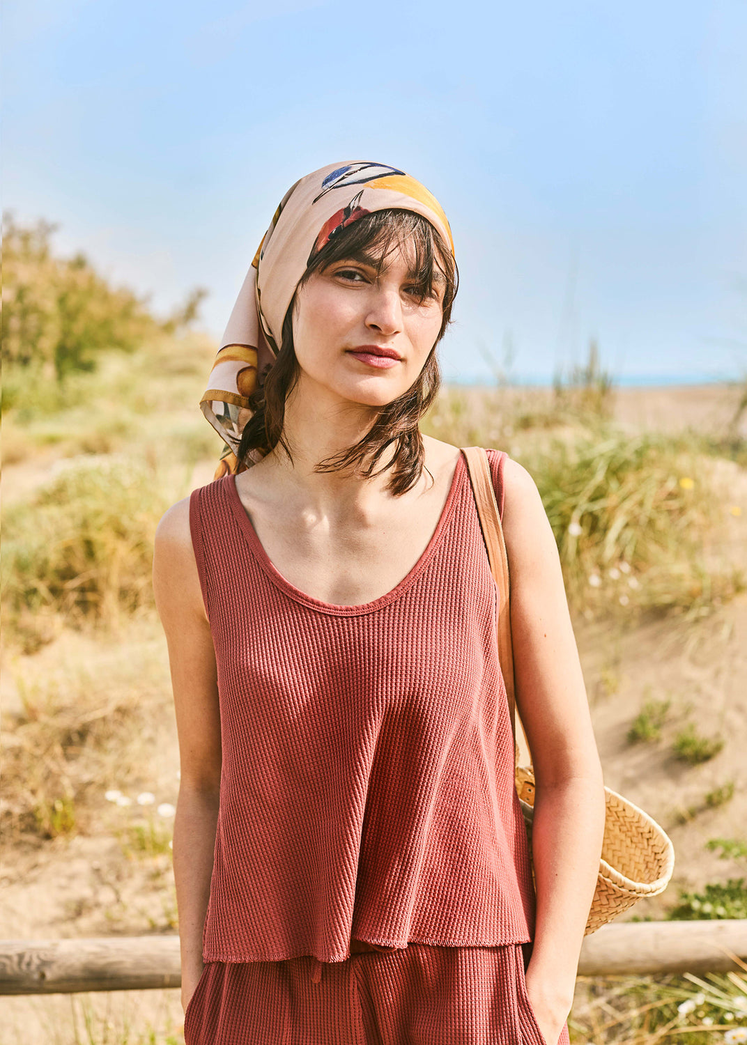 Woman wearing a robin print square scarf tied over her hair, styled with a textured summer set in a Mediterranean coastal setting.